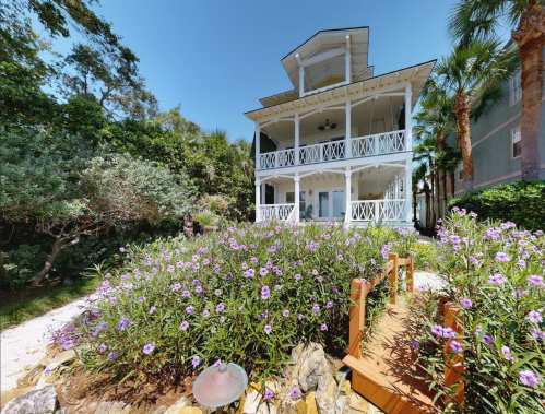 A charming white house surrounded by lush greenery and vibrant purple flowers under a clear blue sky.