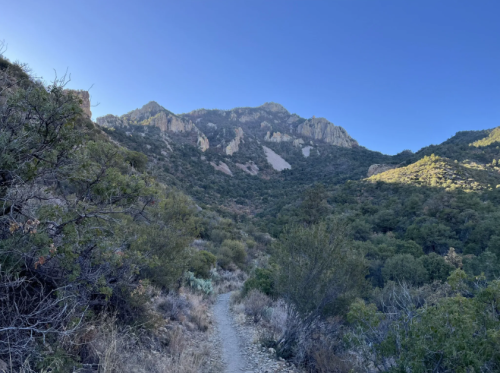 A winding trail leads through a lush valley, surrounded by mountains under a clear blue sky.