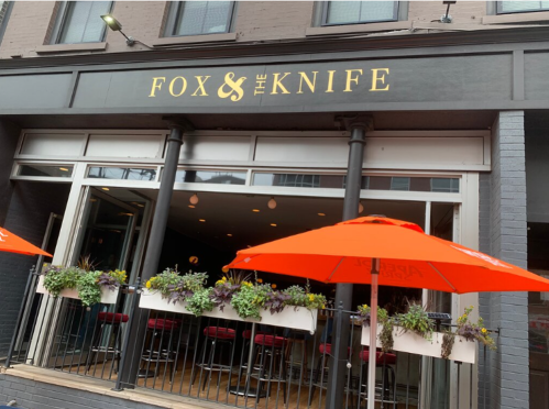 Exterior of "Fox & The Knife" restaurant with orange umbrellas and planters on a balcony.