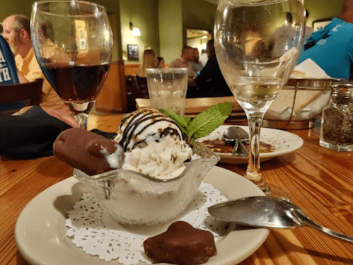 A dessert bowl with ice cream, chocolate-covered treats, and mint, surrounded by glasses of wine in a restaurant setting.