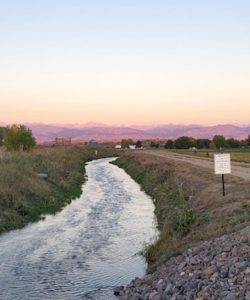A winding river flows through grassy terrain, with mountains in the background and a pastel sky at sunset.