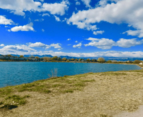 A serene lake surrounded by grassy banks, with mountains and a blue sky filled with fluffy clouds in the background.