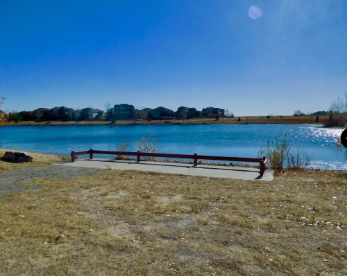 A serene lake view with a wooden railing, surrounded by dry grass and distant houses under a clear blue sky.