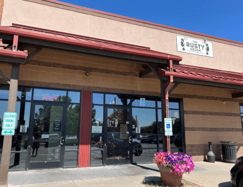 Exterior of The Rusty Melon restaurant, featuring large glass doors and colorful flower pots under a clear blue sky.