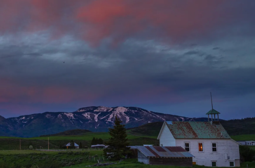 A rustic white building with a green roof against a backdrop of mountains and a colorful sunset sky.