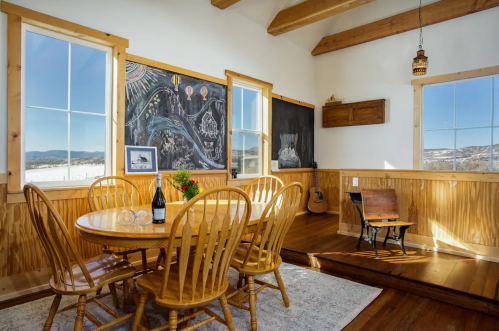 Cozy dining area with a wooden table, chairs, chalkboard wall, and large windows showcasing a scenic view.