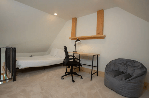 Cozy attic room with a bed, desk, chair, and bean bag, featuring soft lighting and neutral decor.