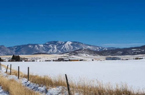 Snow-covered landscape with a mountain in the background under a clear blue sky, featuring a few buildings and a fence.