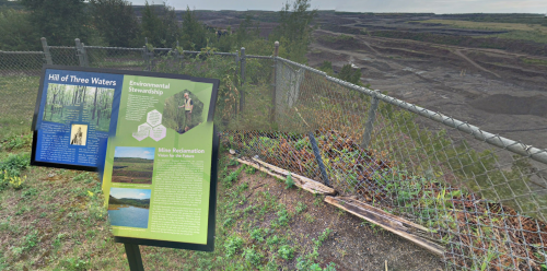 Sign about environmental stewardship and mine reclamation near a large, open-pit mining area with trees in the background.