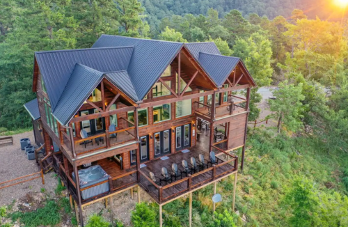 A large wooden cabin with a modern design, surrounded by trees and mountains, featuring multiple balconies and a hot tub.