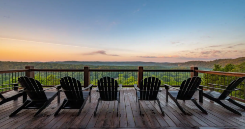 A serene view from a deck with five Adirondack chairs overlooking a lush green valley at sunset.