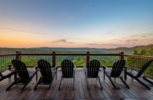 A serene deck with five Adirondack chairs overlooking a lush valley at sunrise.
