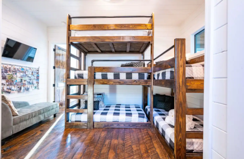 A cozy room with two wooden bunk beds, checkered bedding, and a sofa, featuring wooden floors and bright windows.