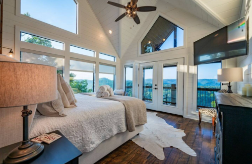 Cozy bedroom with large windows, mountain views, a king bed, and modern decor, featuring a ceiling fan and TV.