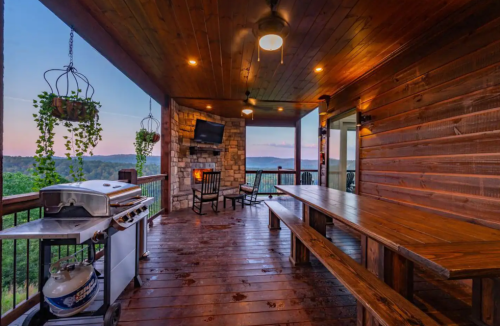 Cozy outdoor patio with wooden furniture, a grill, and a stone fireplace, overlooking a scenic sunset view.