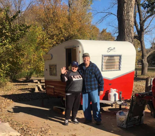 A couple stands smiling in front of a vintage camper surrounded by autumn trees.