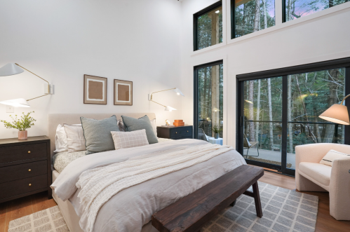 Cozy bedroom with a large bed, modern decor, and windows overlooking a forested area. Warm lighting and neutral tones.