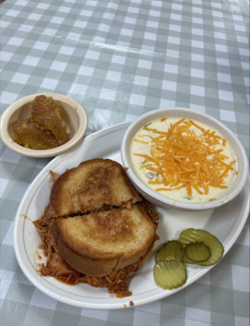 A plate with a toasted sandwich, creamy soup topped with cheese, pickles, and a small dish of dessert.