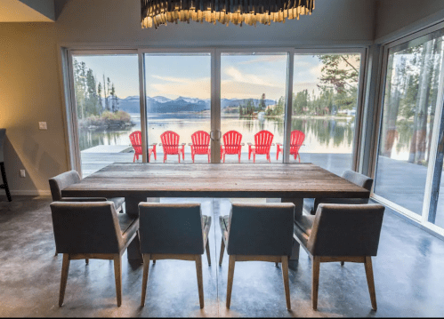 A modern dining area with a wooden table and chairs, overlooking a lake with red chairs on the deck outside.