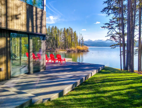 Modern cabin by a serene lake, featuring red chairs on a patio, surrounded by trees and mountains in the background.