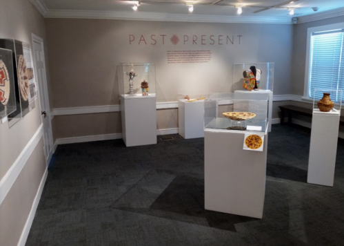Art gallery featuring displays of pottery and artifacts under the theme "Past Present." Brightly lit, neutral walls.