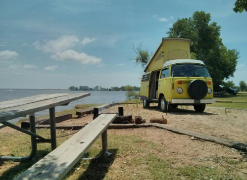 A yellow vintage camper van parked by a lake, with picnic tables and trees in the background under a clear blue sky.