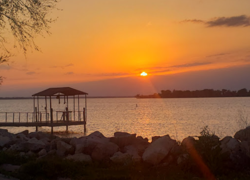 Sunset over a calm lake, with a silhouette of a dock and rocks in the foreground. Soft orange and purple hues in the sky.