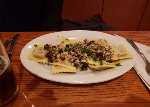 Plate of ravioli topped with chopped nuts and herbs, served on a wooden table with utensils nearby.