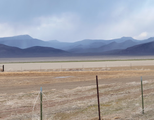A vast, open landscape with distant mountains under a cloudy sky, featuring a barbed wire fence in the foreground.