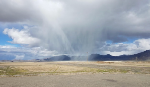 A dramatic sky with dark clouds and rain falling over a barren landscape and distant mountains.