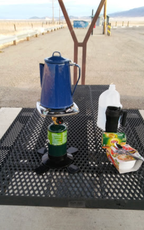 A blue kettle on a camping stove, with a water jug, cup, and instant noodles on a picnic table.