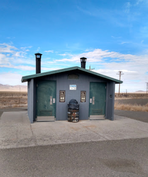 A small, gray restroom building with two doors, surrounded by open land and a clear blue sky.