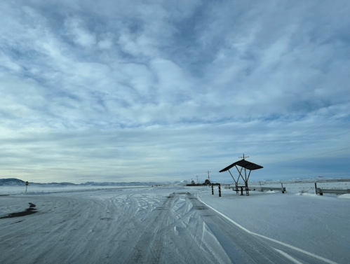 A snowy landscape with a cloudy sky, featuring a shelter and road signs in the distance.