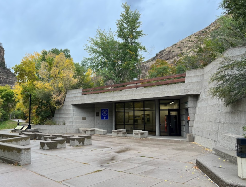A modern building with a stone facade, surrounded by trees and rocky hills, under a cloudy sky.