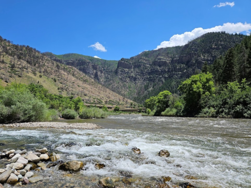 A scenic river flows through a valley surrounded by mountains and lush greenery under a clear blue sky.