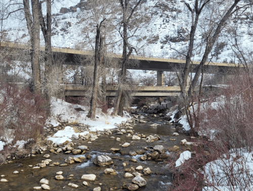 A snowy landscape featuring a stream with rocks, flanked by trees and an overpass above.