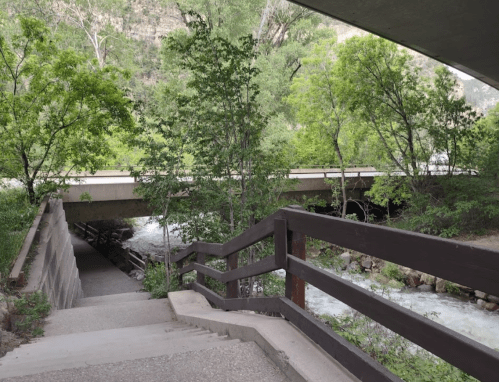 A pathway with wooden railing leads down to a river, with a bridge and lush greenery in the background.