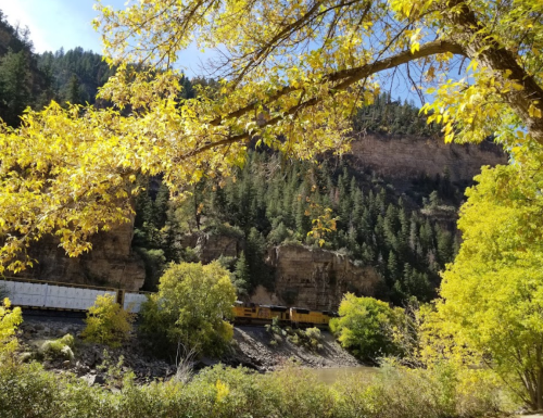 A train travels along a river surrounded by vibrant autumn foliage and rocky cliffs under a clear blue sky.