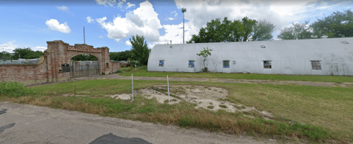 An old brick archway leads to a white, curved building surrounded by grass and trees under a partly cloudy sky.