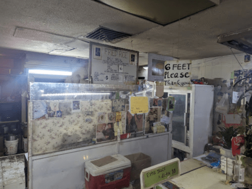 A small, cluttered deli with a sign for fried rice and a reminder to maintain social distance.