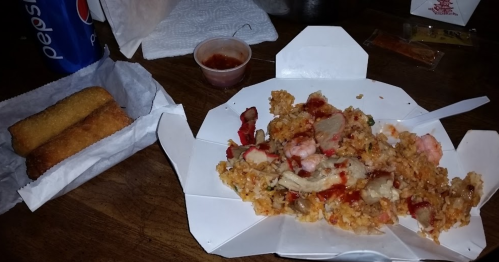 A takeout box with fried rice topped with meat and sauce, alongside a container of dipping sauce and spring rolls.