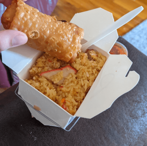 A hand holding an egg roll above a takeout box of fried rice with pieces of meat and a small container of sauce.