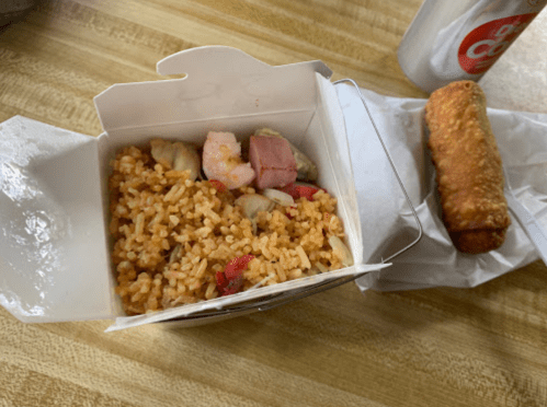 Takeout box with fried rice and shrimp, alongside a spring roll and a can of soda on a wooden table.