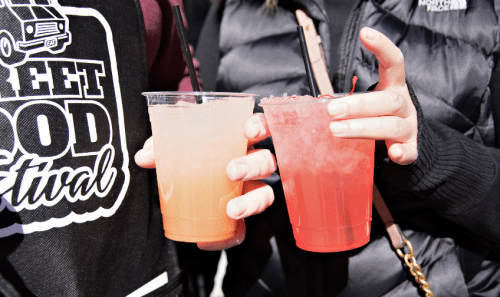 Two hands hold colorful drinks with straws at a street food festival, one orange and one red.
