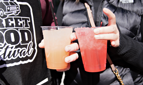 Two hands hold colorful drinks with straws at a street food festival, one orange and one red.