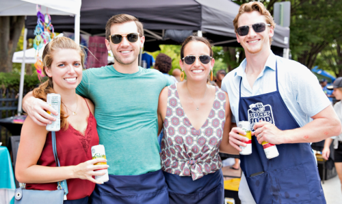 Four friends smiling at an outdoor event, holding drinks and wearing sunglasses, with tents in the background.