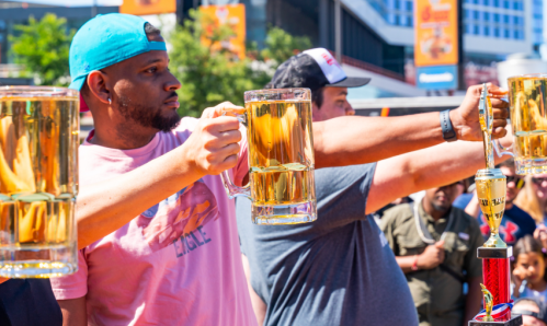 Two men hold up large mugs of beer in a competition, with a trophy visible in the foreground. Bright outdoor setting.
