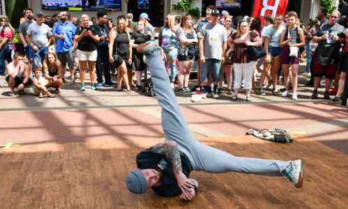 A breakdancer performs on a wooden floor while a crowd watches in an outdoor setting.