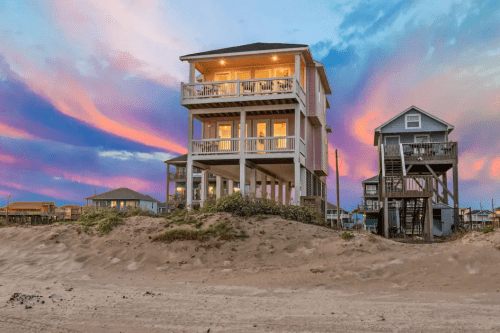 A beach house with multiple levels stands against a colorful sunset sky, surrounded by sand and other homes.