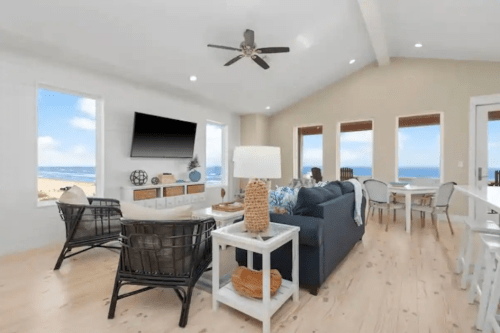 Bright coastal living room with ocean views, featuring a sofa, chairs, and a dining area with large windows.